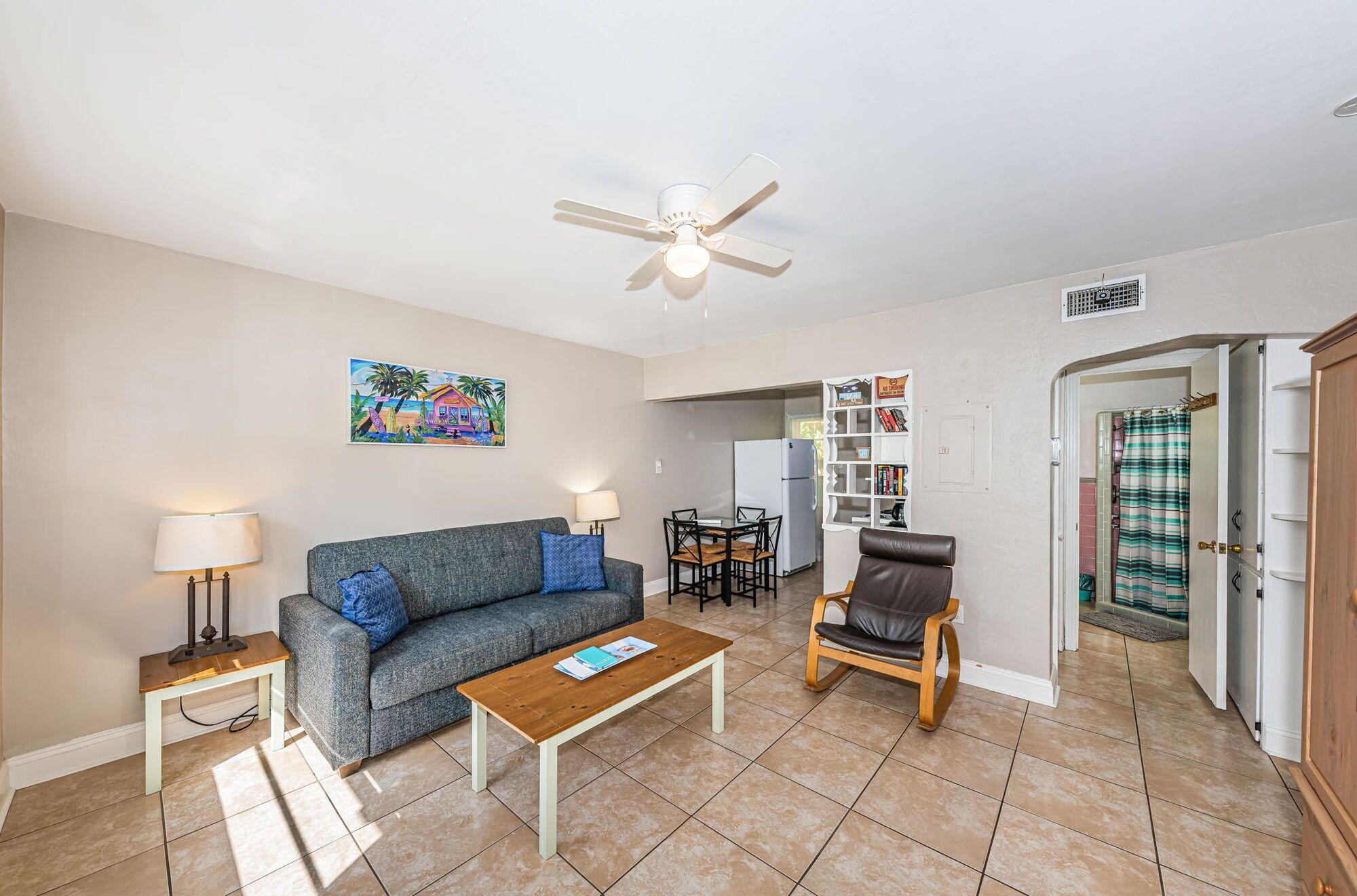 A living room with a sofa, coffee table, and a chair. There is a ceiling fan, a painting on the wall, and a kitchen area visible through an open door.