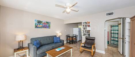 A living room with a sofa, coffee table, and a chair. There is a ceiling fan, a painting on the wall, and a kitchen area visible through an open door.