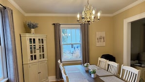 A dining room with a wooden table, white chairs, a chandelier, and a window with curtains. - "The Owl's Nest" Updated Craftsman home overlooking Old Town Park. (Westerville)