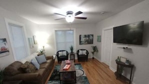 A living room with a brown sofa, two black chairs, a coffee table, a ceiling fan, a TV on the wall, and a wooden floor. - Beachcomber! Pro Cleaned, Self Check in Sleeps 7 (Racine)