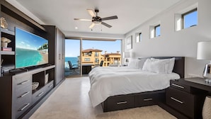 Une chambre moderne avec un grand lit, un ventilateur de plafond, un téléviseur posé sur un meuble sombre et une vue sur l’océan par une porte-fenêtre coulissante.