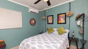 Un dormitorio con cama, ventilador de techo, aire acondicionado, mesita de noche, lámpara y perchero en la pared.