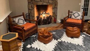 A cozy living room with a stone fireplace, two wooden armchairs with cushions, a small wooden table, and a leather pouf. - BigBear Lodge: Mt. Sunapee / Hot tub / Firepit (New London)