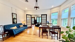 Ein großzügiger Wohnraum mit blauem Sofa, Esstisch aus Holz und Stühlen, großen Fenstern und einer Kristallleuchter.
