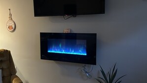 Sala de estar con un televisor de pantalla plana montado en la pared, una chimenea debajo y suelo de madera, además de una silla.