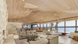 Amplia sala de estar con un ventanal que da a una masa de agua, techos altos con vigas de madera, pared de acento de piedra y muebles modernos.