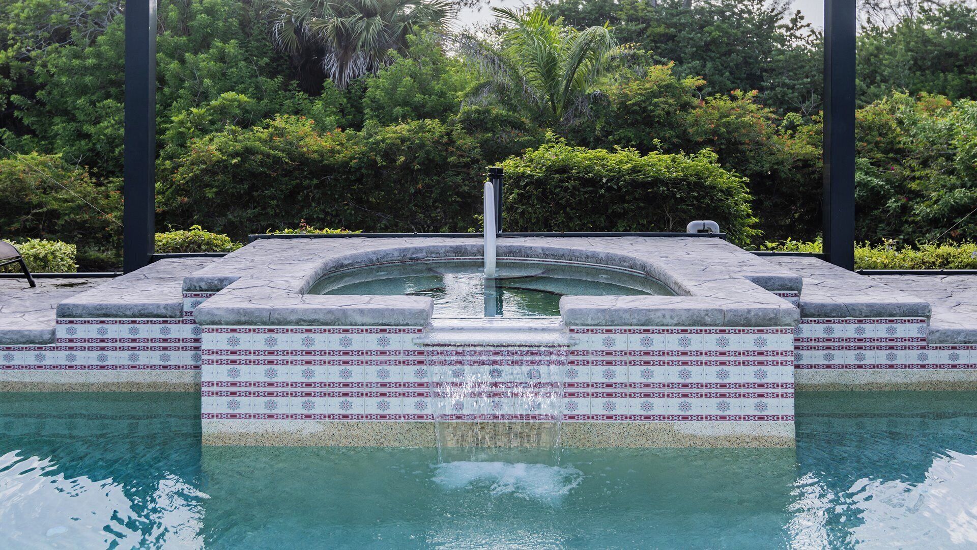 A swimming pool with a fountain, surrounded by a tiled border and lush greenery.