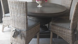 Een eetruimte met een ronde tafel, vier rieten stoelen, een vaas met roze bloemen en een spiegel aan de muur.