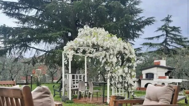 Una terraza de madera con dos sillas y una mesa, un arco blanco con flores y un edificio al fondo.