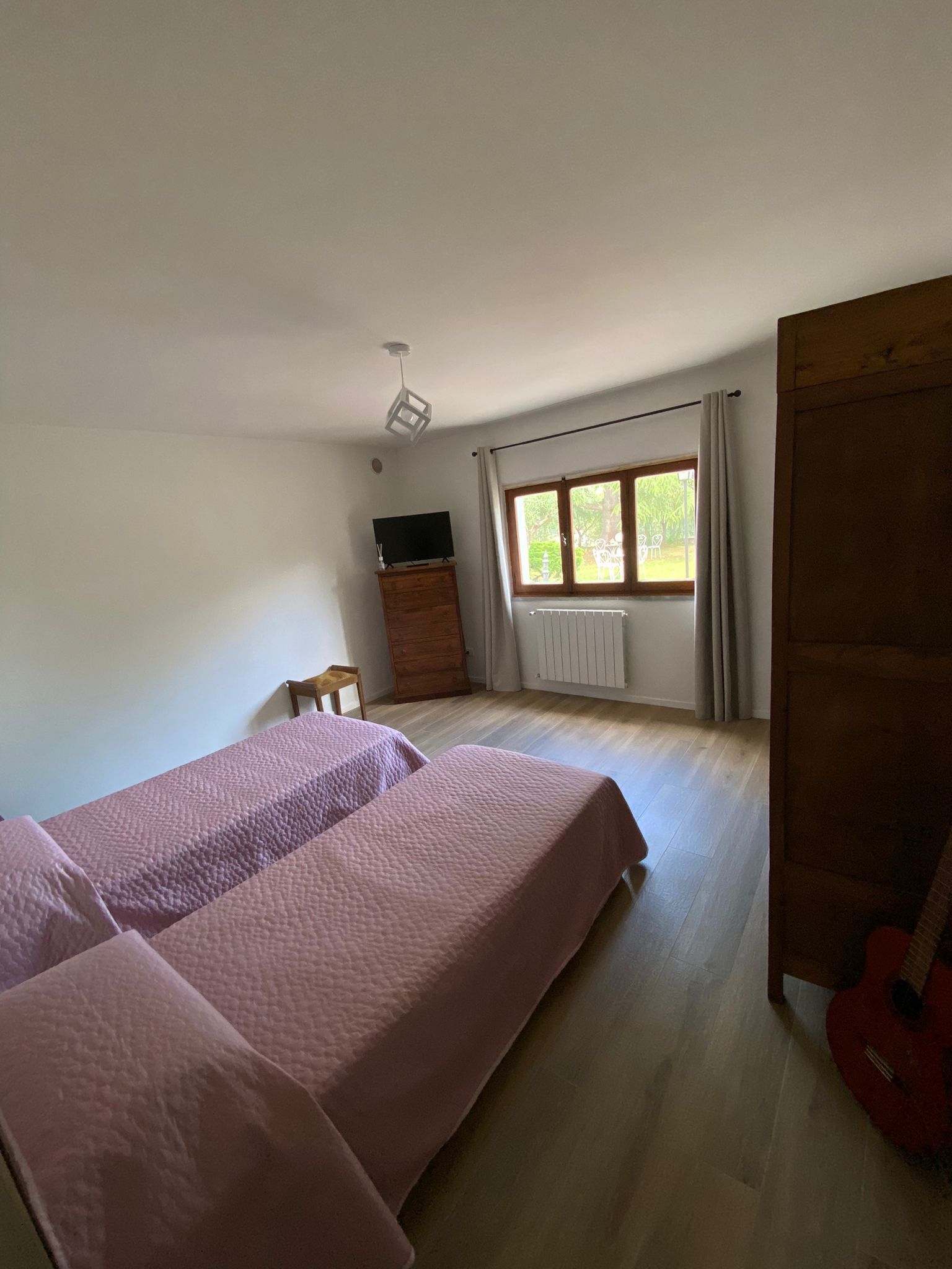 Een slaapkamer met twee bedden, een houten dressoir, een televisie, een raam met gordijnen en een gitaar.
