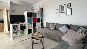 Een moderne woonkamer met een grijze bank, een klein zwart tafeltje en een tv op een witte dressoir. Er staan kleurrijke opbergkasten en er is een plafondventilator.