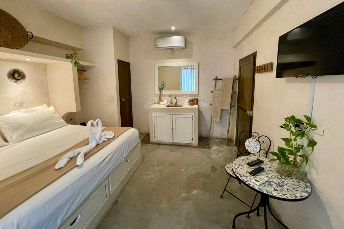 A hotel room with a bed, bedside table, a small round table with a chair, a TV mounted on the wall, and a bathroom with a sink and mirror.
