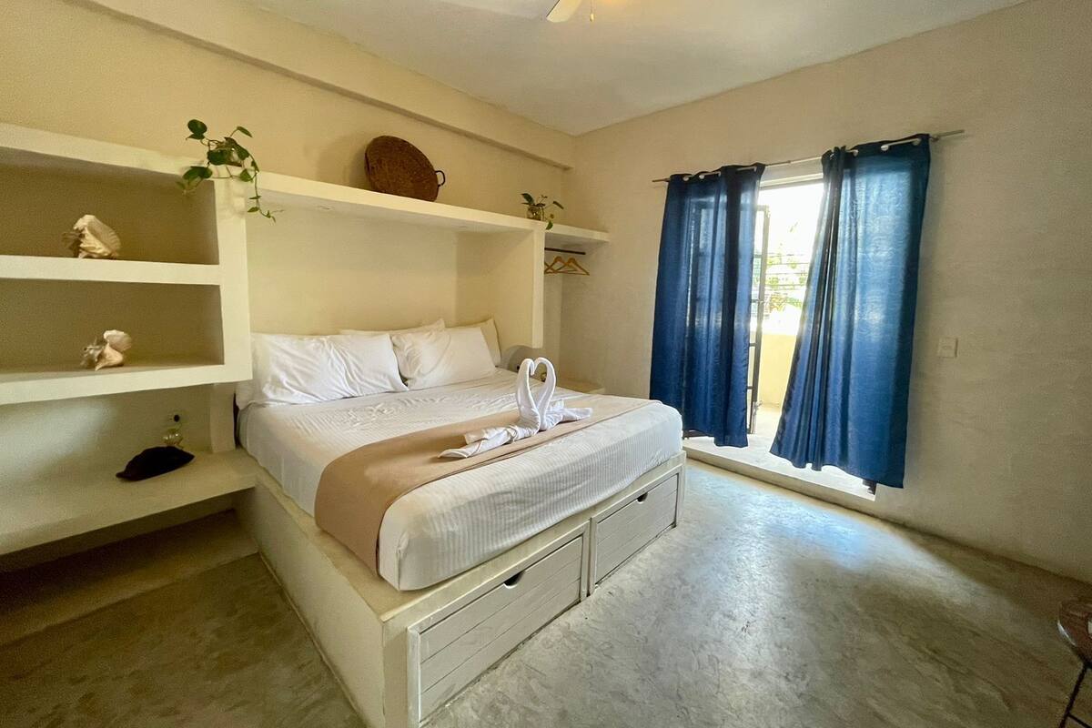 A bedroom with a bed, a ceiling fan, a window with curtains, and a shelf with decorative items.