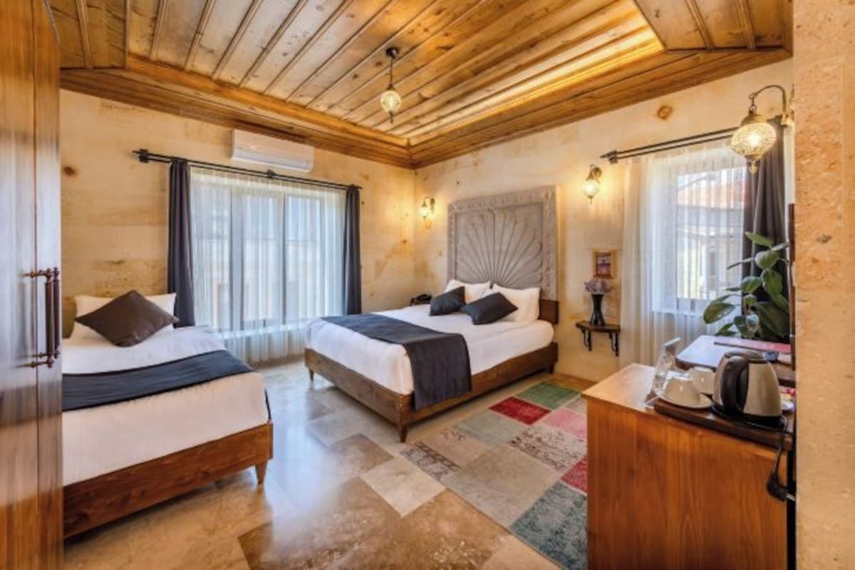 Habitación de hotel con dos camas, suelo y techo de madera, un escritorio pequeño con hervidor y una ventana con cortinas.