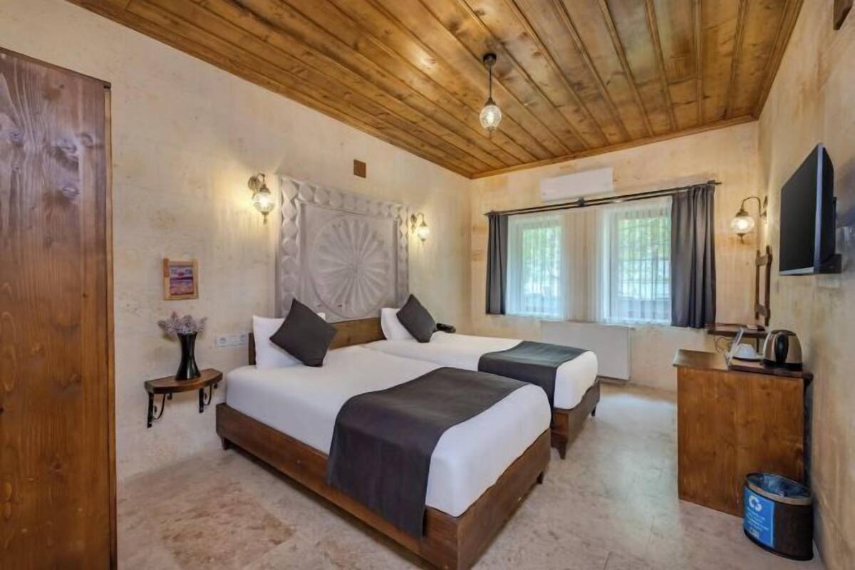 A room with two beds, wooden ceiling, wall-mounted light fixtures, a television, and a window with curtains.