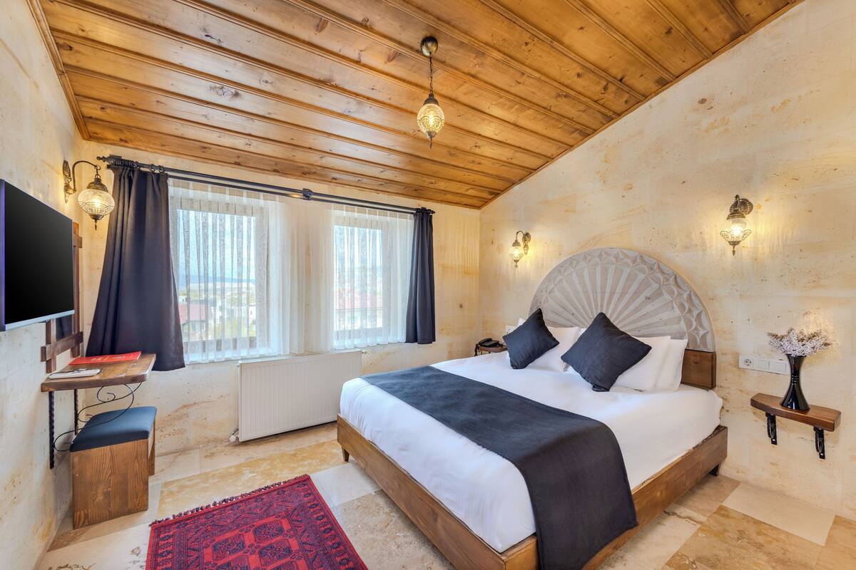 A bedroom with a wooden ceiling, a bed with a headboard, bedside tables, a TV, and a window with curtains.