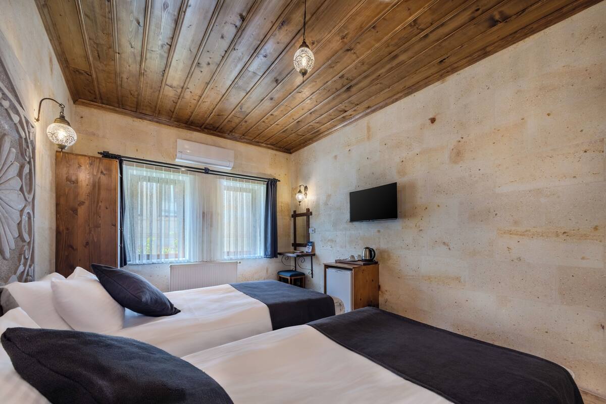 A room with wooden ceiling beams, two beds with white and black bedding, a small table with a kettle, a television on the wall, and a window with curtains.