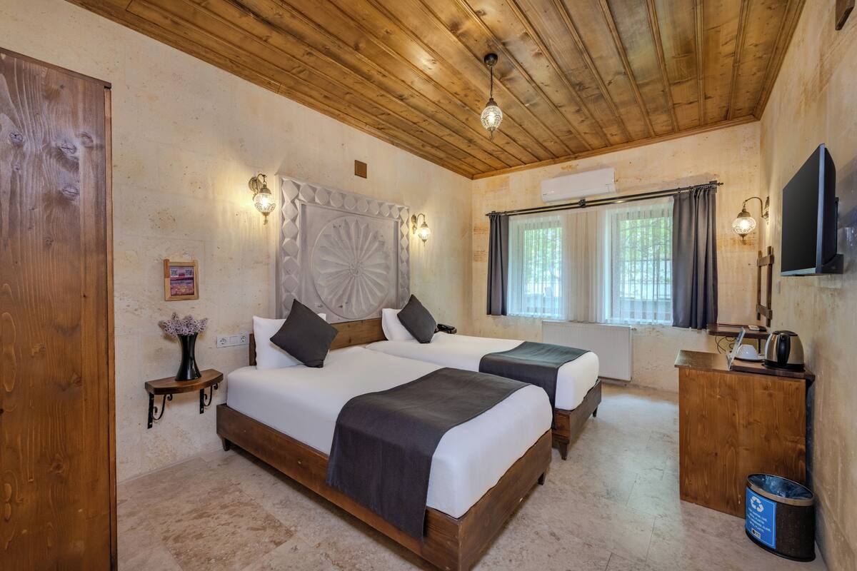 A room with two beds, wooden ceiling, wall-mounted TV, and a small table with a vase.