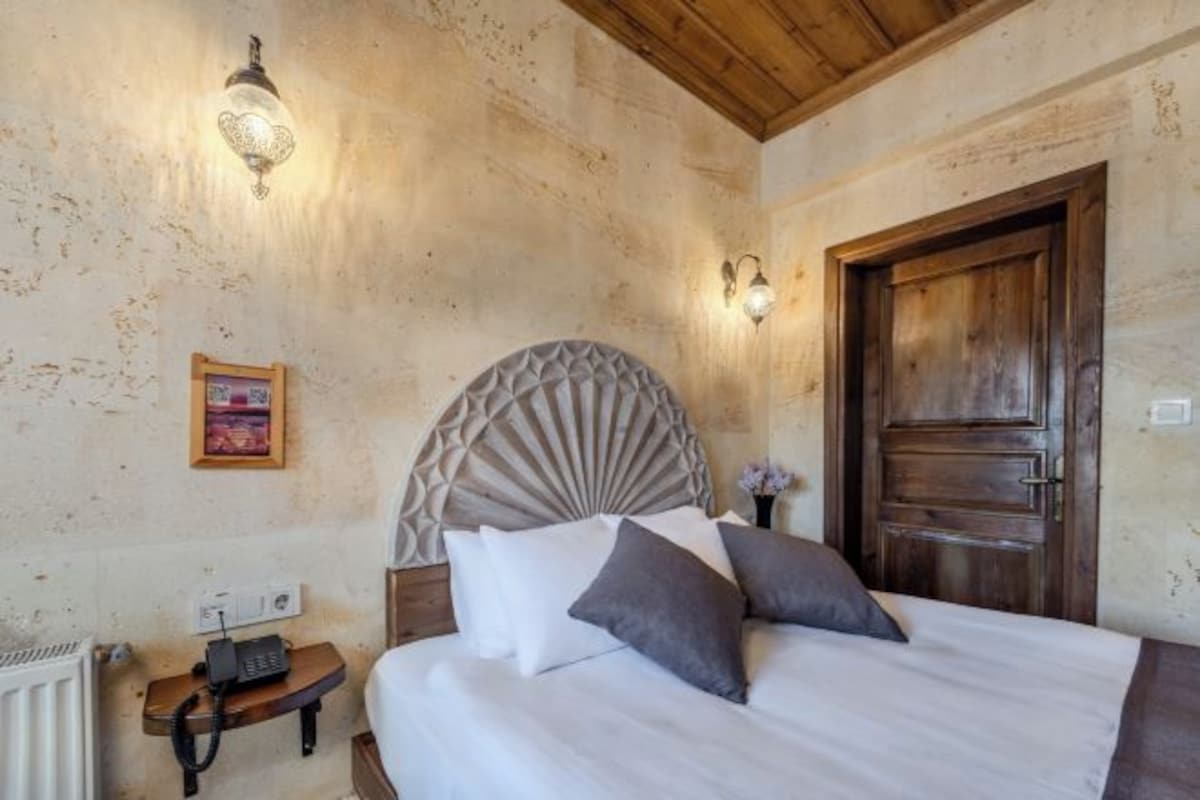 Un dormitorio con techo de madera, una cama con ropa de cama blanca y gris, una puerta de madera y dos lámparas fijadas a la pared.