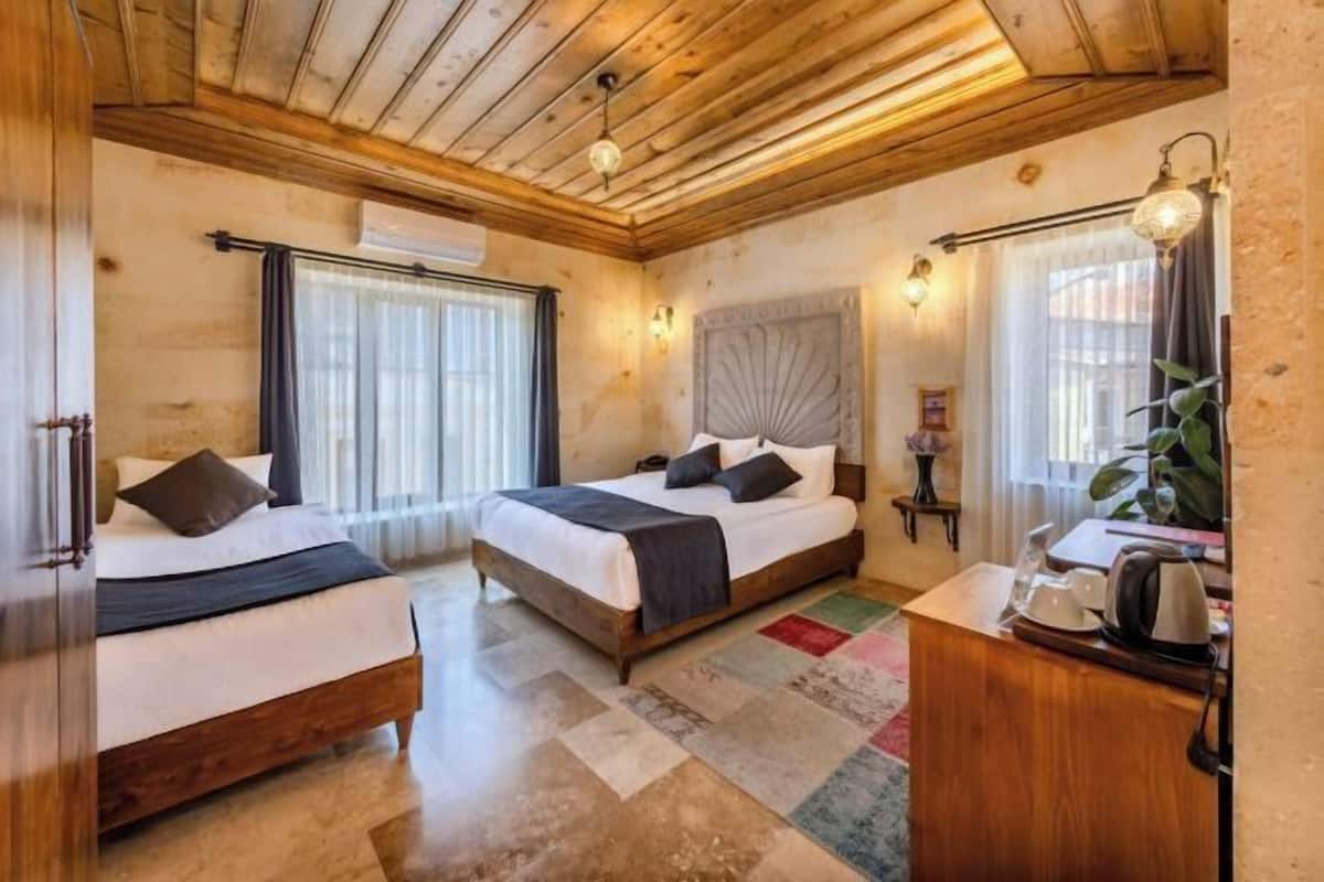 A hotel room with two beds, wooden flooring, a wooden ceiling, a small desk with a kettle, and a window with curtains.
