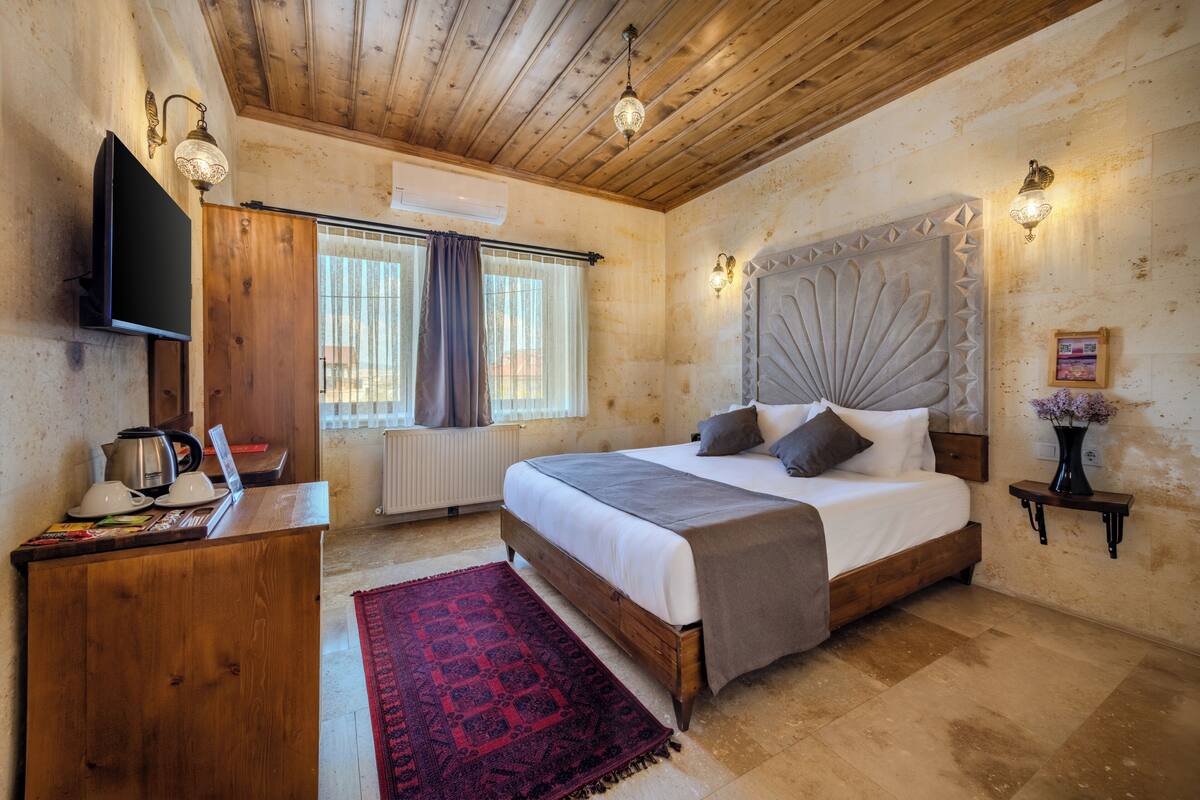 A bedroom with a wooden ceiling, a bed with a headboard, a TV mounted on the wall, and a window with curtains.