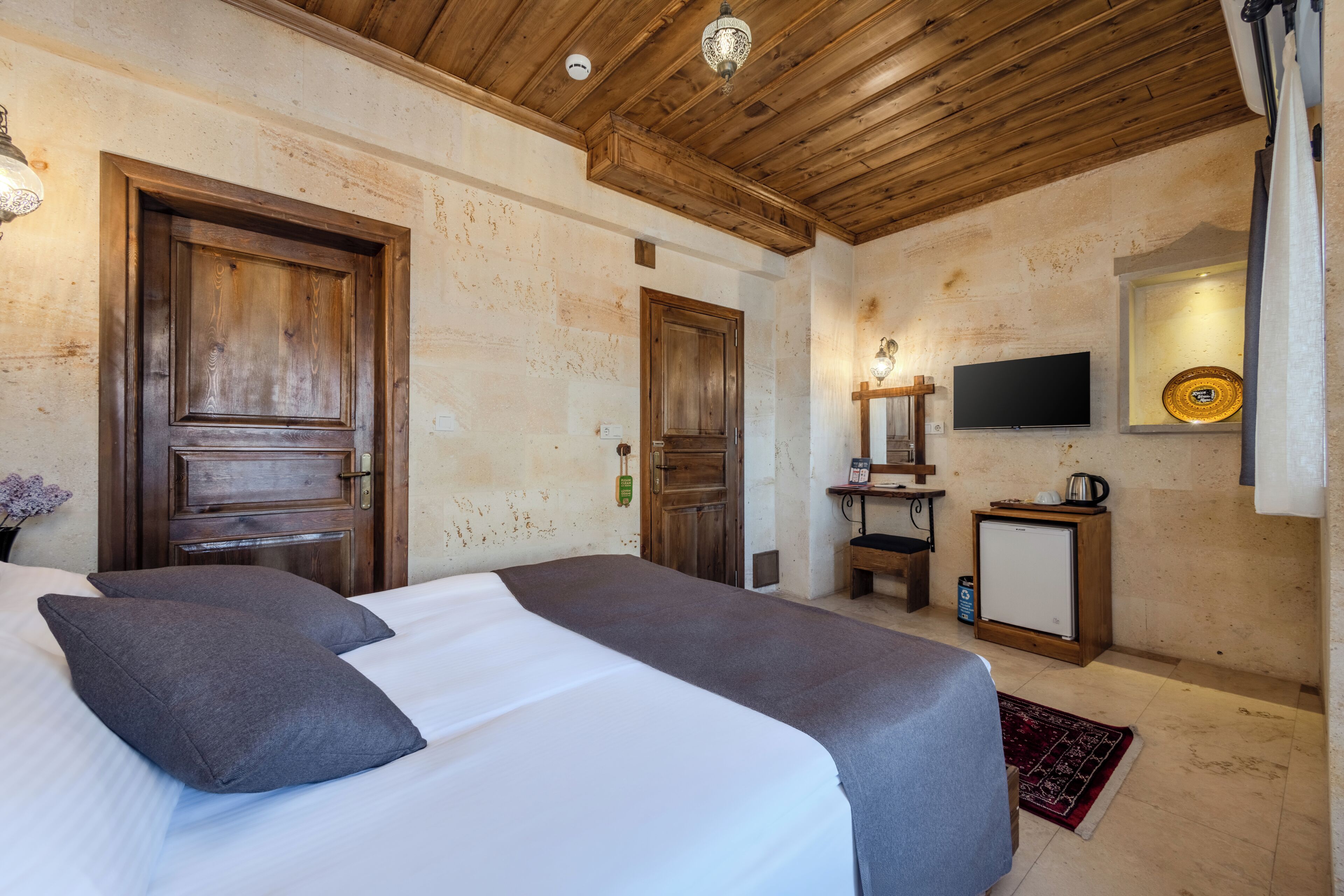 Habitación de hotel con techo de madera, cama con ropa de cama gris, televisor, mesita con tetera y televisor montado en la pared.