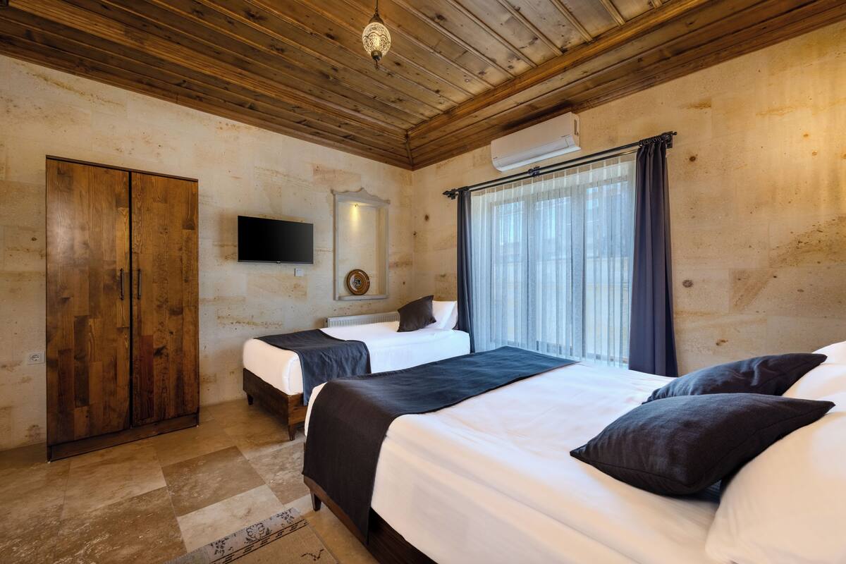 A room with wooden ceiling, two beds with dark bedding, a TV on the wall, and a window with curtains.