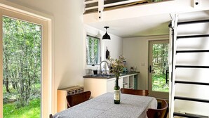 Comedor con una mesa dispuesta para cuatro, cocina al fondo y vista de un bosque a través de un ventanal grande.