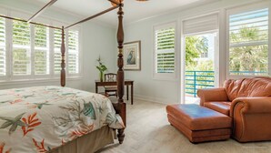 A four-poster bed with a floral bedspread, a leather armchair, and a dining table with chairs in a bright room with large windows and a view of greenery. - Banana Key (Key West)