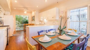 Un coin repas avec une table en bois dressée pour quatre personnes, entourée de chaises. On y trouve une fenêtre avec des stores, un ventilateur de plafond et une vue sur la verdure à l’extérieur.