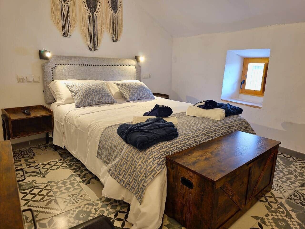 Una camera da letto con un letto, comodini, una panca in legno, un pannello macramè appeso al muro e una piccola finestra.