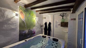 A bathroom with a large bathtub, a shower area, and a wall-mounted heater. There are towels hanging on hooks and a small shelf with a plant.