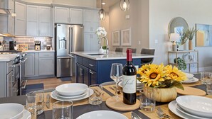A modern kitchen with grey cabinets, a stainless steel refrigerator, and a dining area set with white plates, glasses, and a bottle of wine. - Elegant Home Near TPC Golf Course and Trails (Loveland)