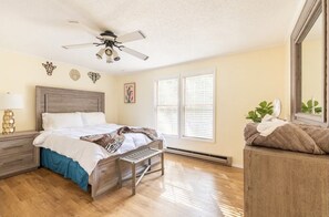 Une chambre à coucher comprenant un lit, une table de chevet, un banc, un ventilateur de plafond et une fenêtre avec des stores.