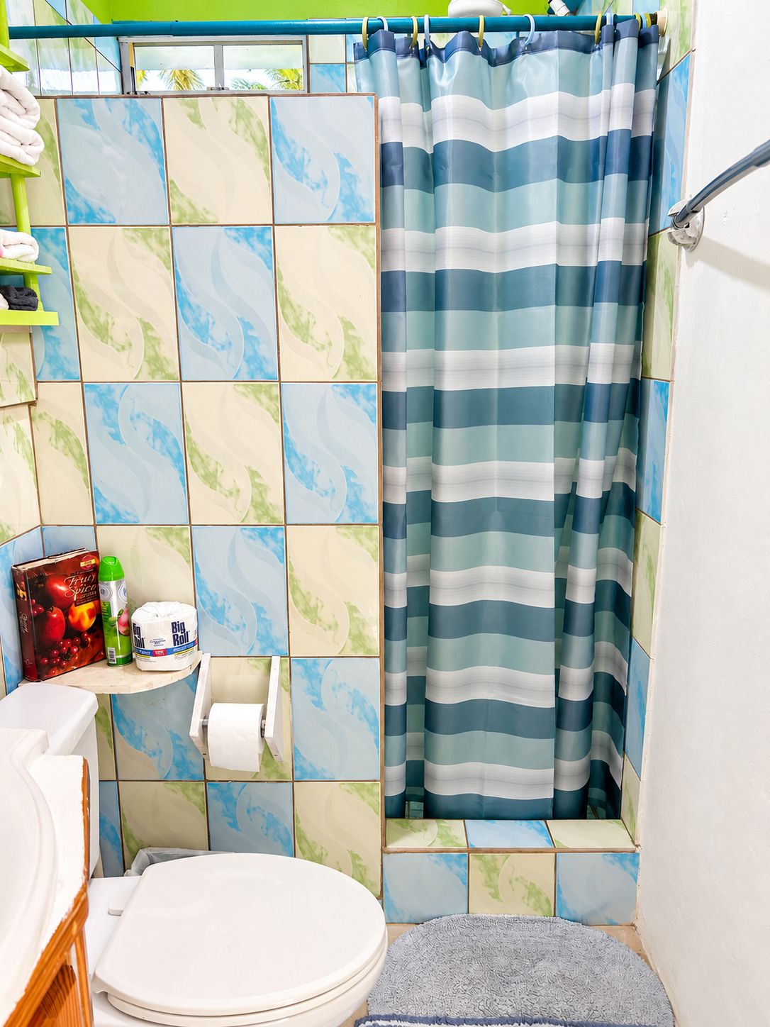 Un baño con inodoro, repisas con artículos de aseo, cortina de ducha y pared con azulejos.