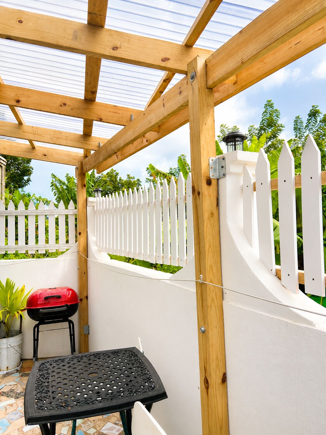 A covered patio area with a wooden pergola, a white picket fence, a red barbecue grill, and a black metal table.