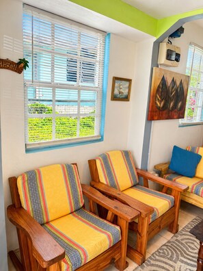 A room with two wooden chairs with yellow cushions, a wooden table, a window with blinds, and a wall-mounted air conditioning unit. - NIZA’S PLACE—Cozy Mountainside Villa with Ocean view. (Paix Bouche)