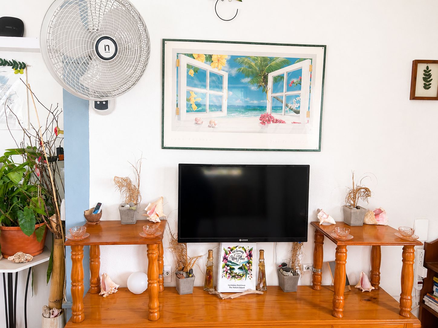 Una sala de estar con un televisor de pantalla plana, dos mesitas de madera, un cuadro enmarco con una escena tropical, un ventilador de pie y plantas en macetas.