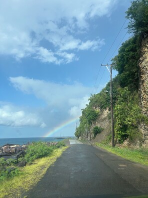 A paved road with a stone wall on one side and a grassy area on the other, leading to a body of water with a rainbow in the distance. - NIZA’S PLACE—Cozy Mountainside Villa with Ocean view. (Paix Bouche)