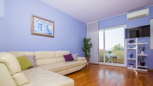 A living room with a beige sofa, a framed painting, a potted plant, and a TV on a white stand. - Apartment 3 beaches with free parking (Dubrovnik)