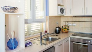 A modern kitchen with white cabinets, a dishwasher, an oven, and a window with blinds. - Apartment 3 beaches with free parking (Dubrovnik)