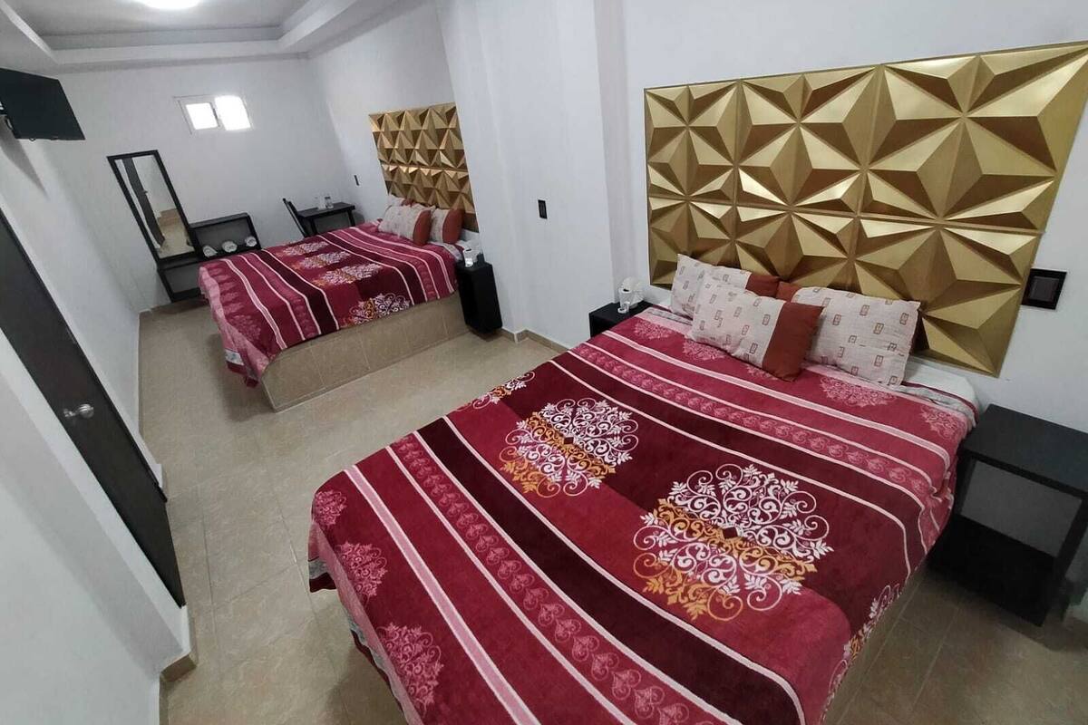 A hotel room with two beds, each with a red and white patterned bedspread, a gold geometric headboard, and a small bedside table with a lamp.