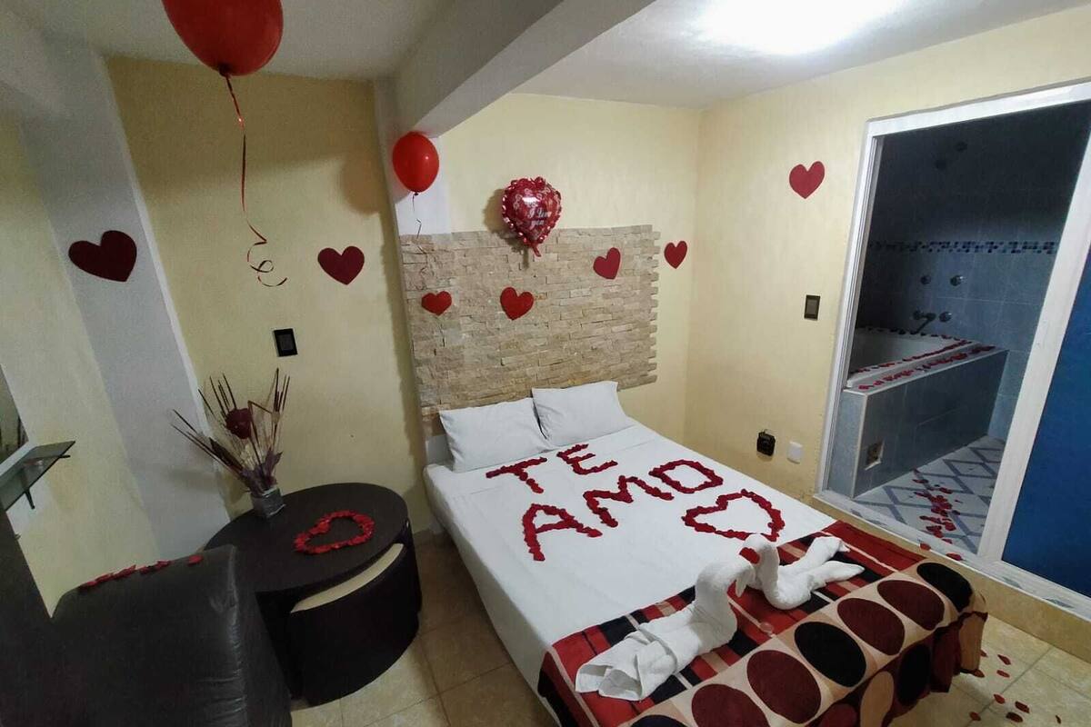 A hotel room with a bed decorated with red heart shapes and the words "TE AMO". There is a small round table with a red heart decoration, a black leather chair, and a glass shower enclosure with rose petals.