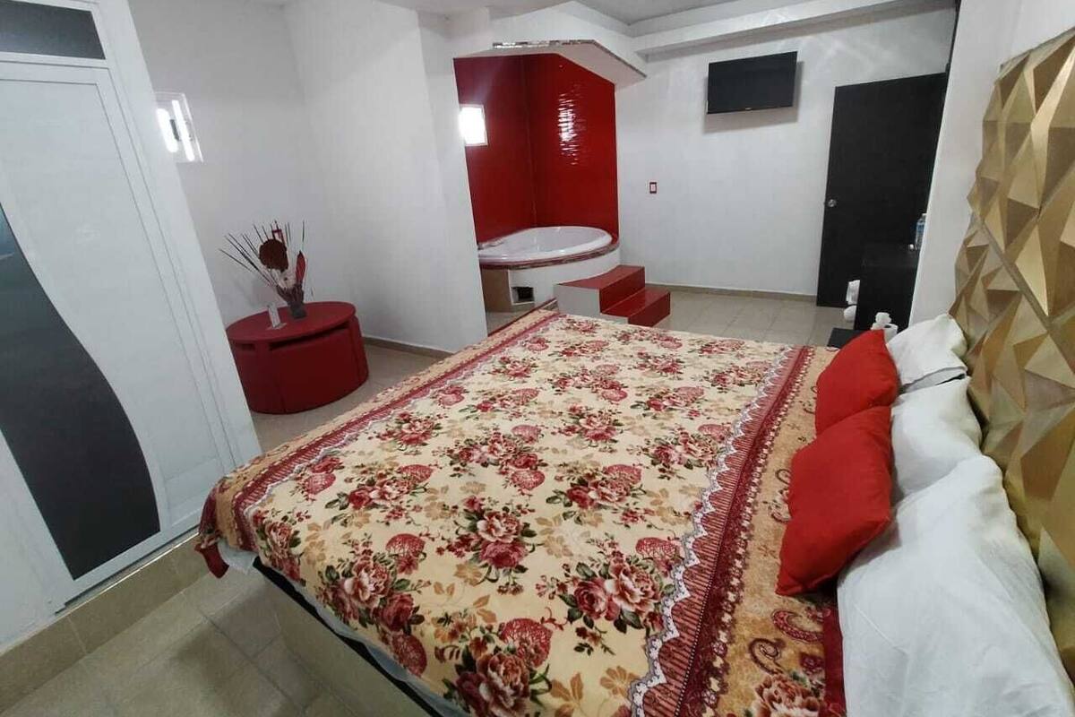 A bedroom with a large bed, a red accent wall, and a small red table with a vase.
