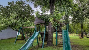 Ein Spielplatz mit zwei Rutschen und einer Kletterkonstruktion, umgeben von Bäumen und Rasen.