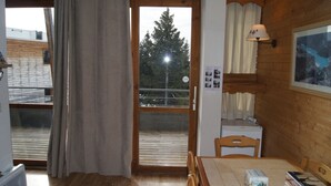 A room with a wooden floor, a table with chairs, a window with curtains, a small white cabinet, and a wall-mounted lamp. - Appartement 6 pers proche pistes (Chamrousse)