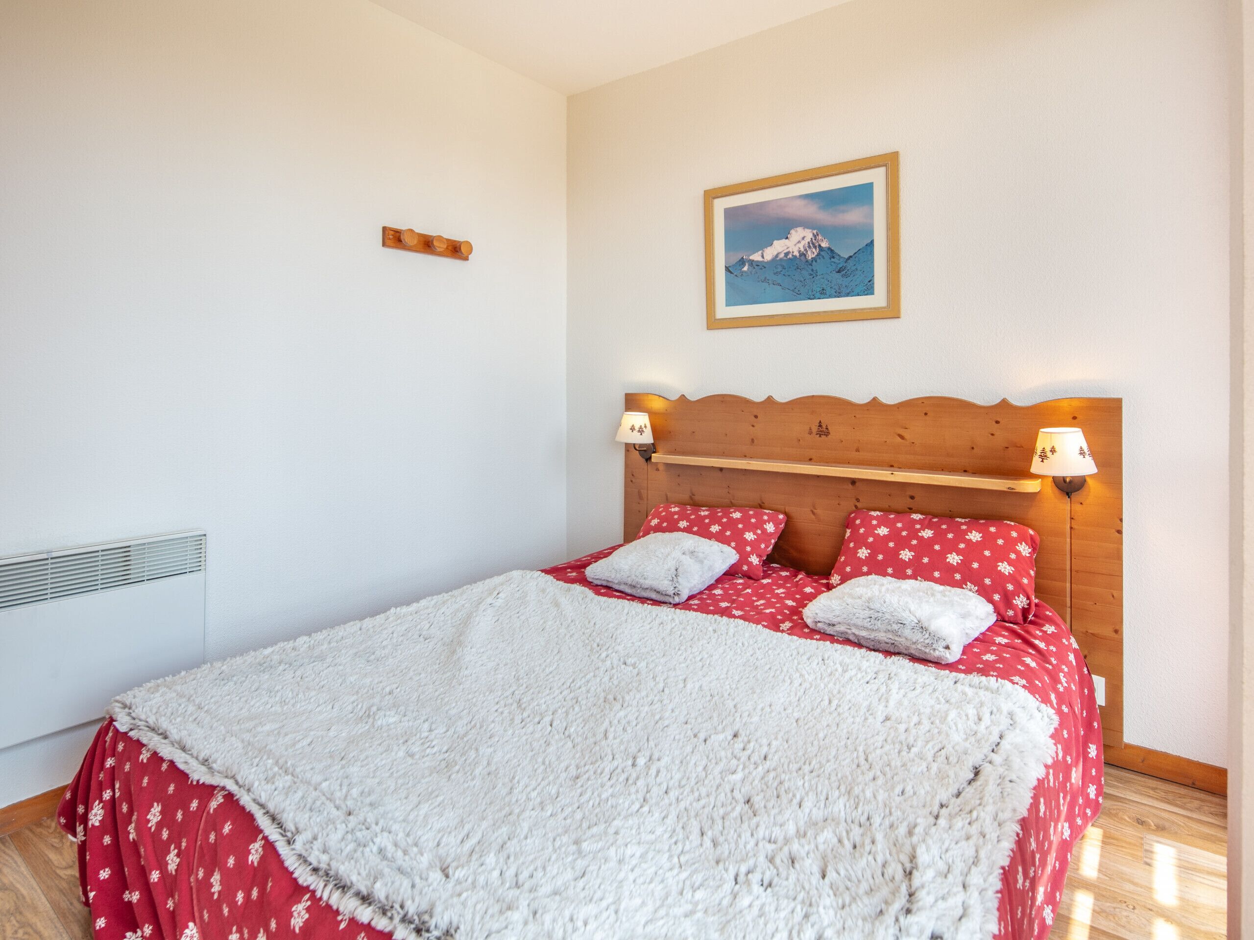 A bedroom with a wooden headboard, two bedside lamps, a framed picture of a mountain, and a red and white patterned bedspread.