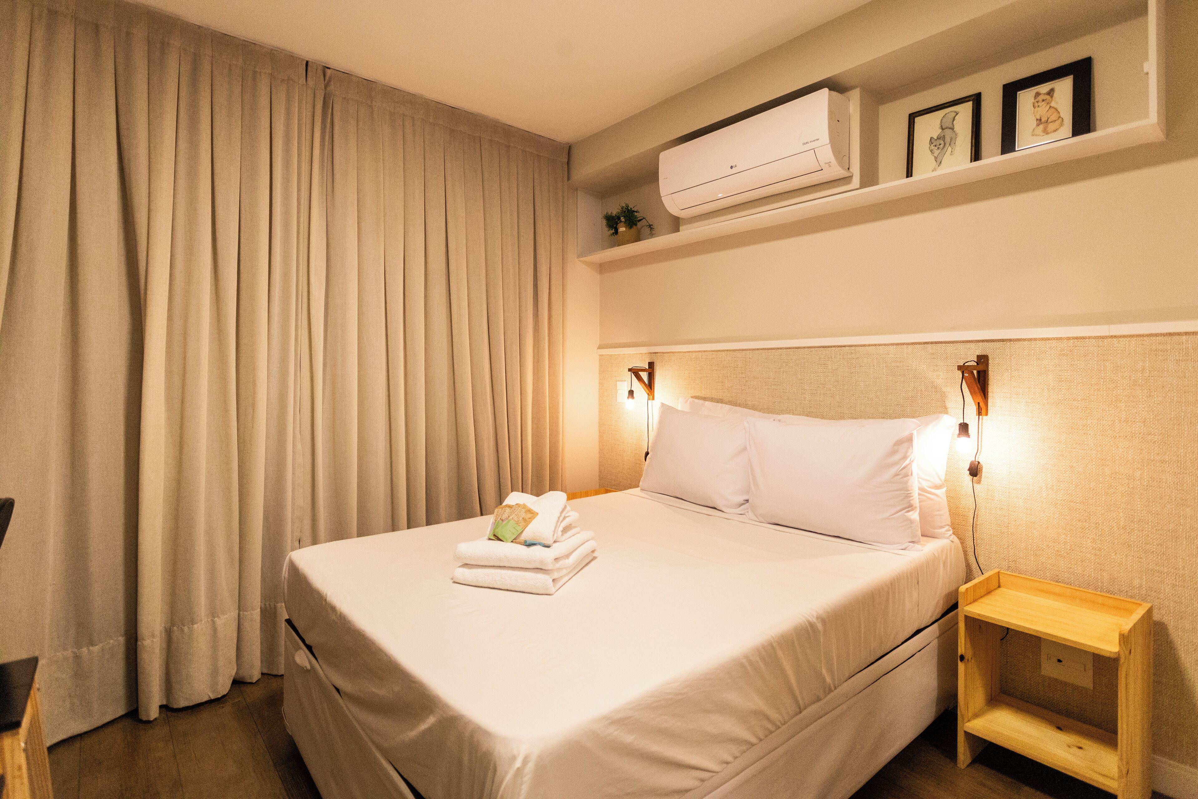 A hotel room with a neatly made bed, a bedside table with a lamp, and an air conditioning unit above the bed.
