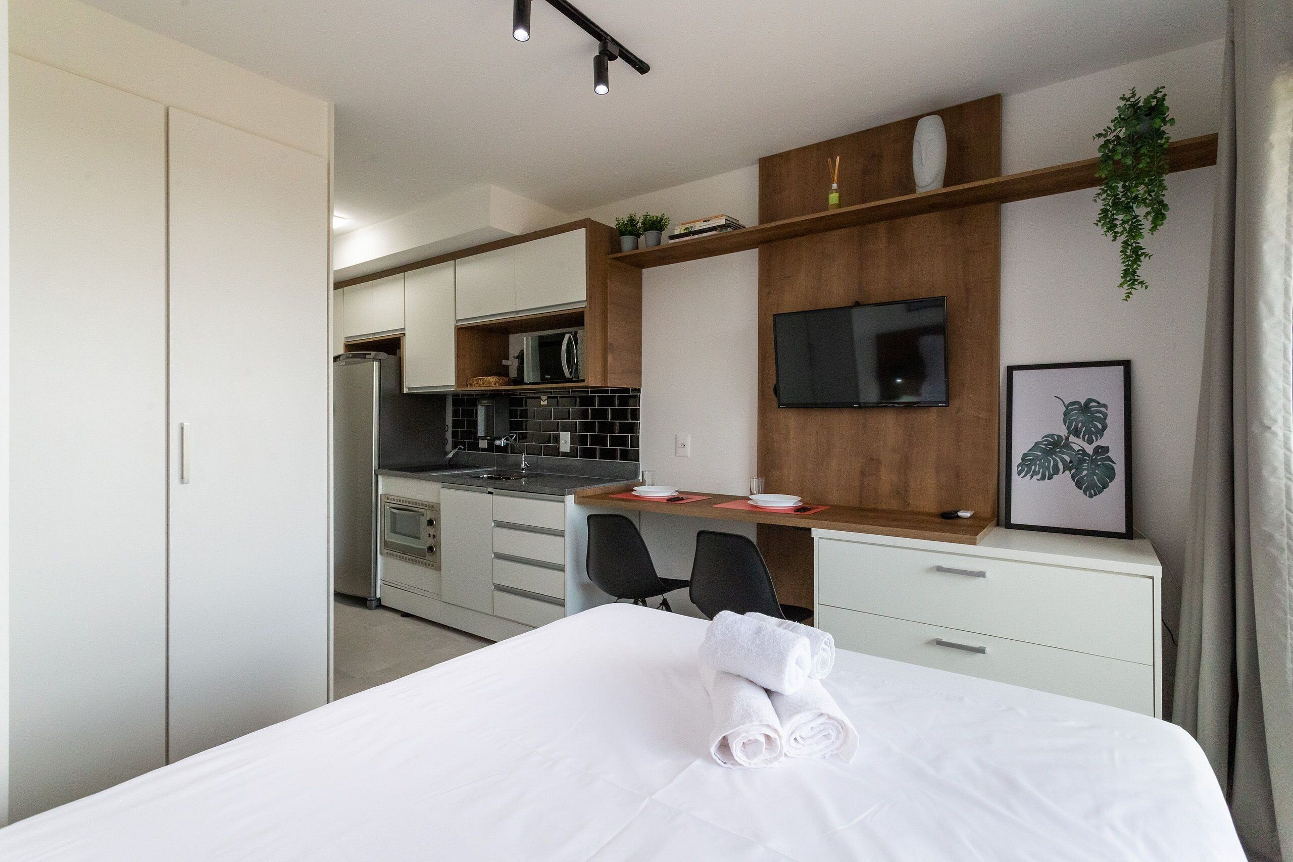 A modern hotel room with a bed, a desk, a TV, and a kitchenette.