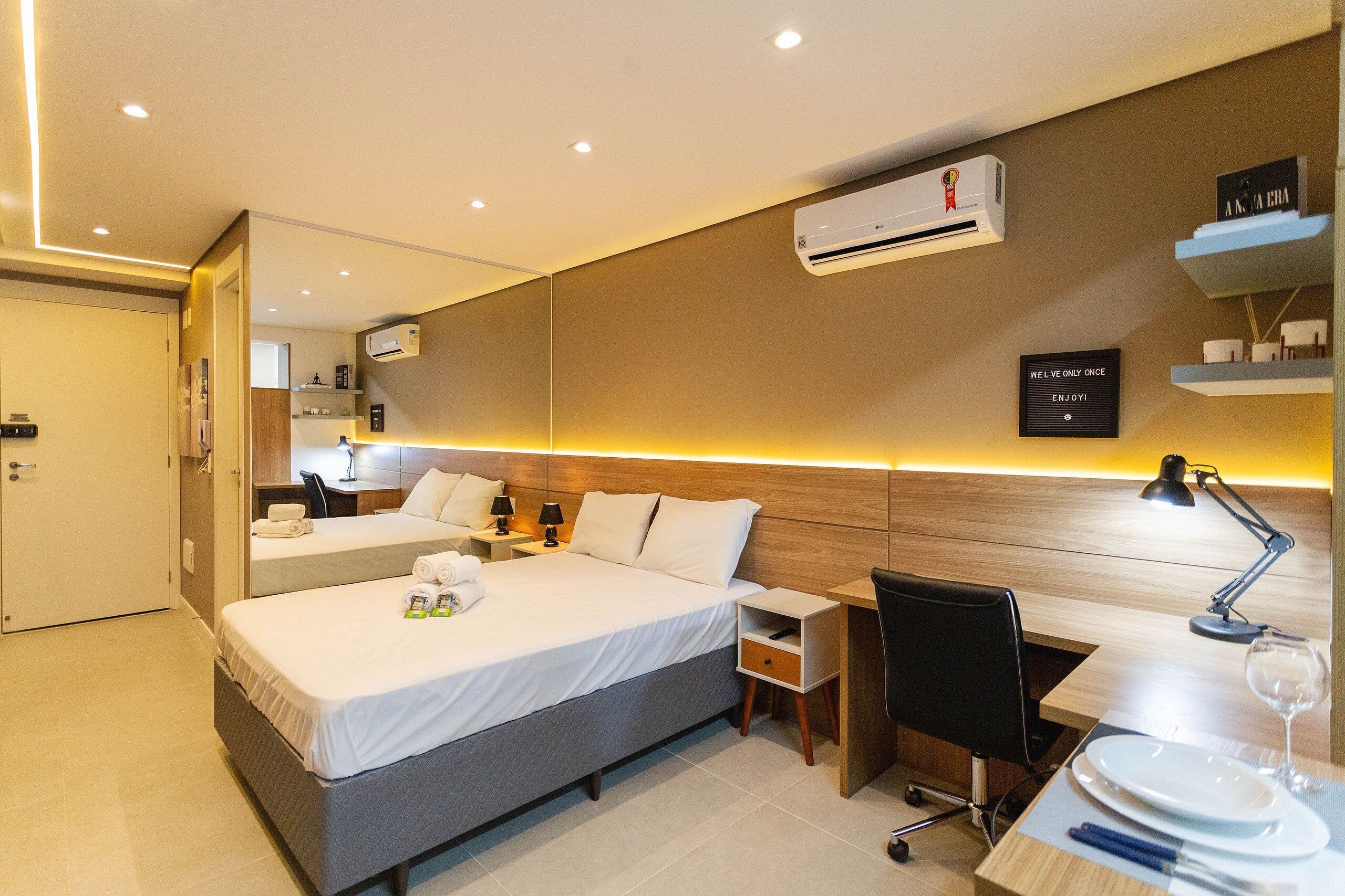A modern hotel room with a bed, desk, chair, and a small dining area with plates and a wine glass.